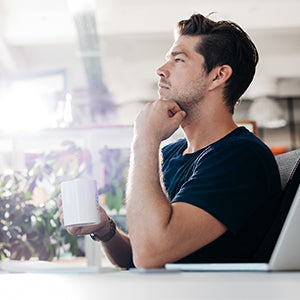 A man with his hand to his jaw, thinking while having a white mug in his other hand.